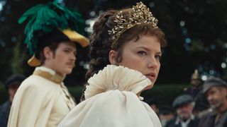 Empress Elisabeth of Austria wearing a crown and looking at something off camera as Emperor Franz stands behind her wearing a feathered hat in The Empress season 2.